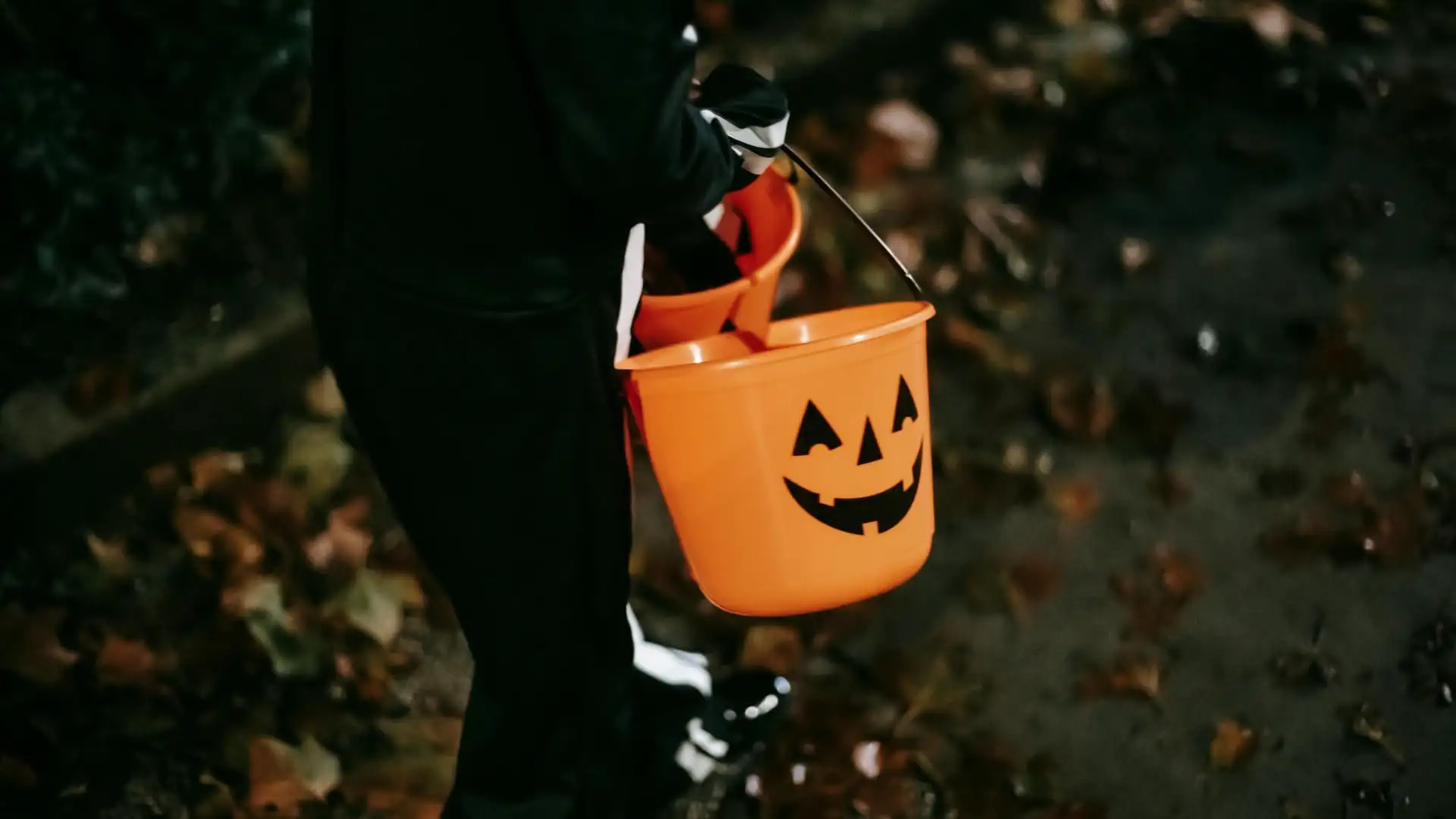 Secchiello arancione a forma di zucca per il dolcetto o scherzetto tenuto da un bambino in costume tra le foglie d’autunno, notte di Halloween | DiBi Group.