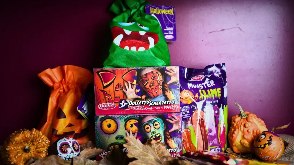 Selezione di dolci Dolfin per Halloween con confezioni a tema, zucche decorative e foglie autunnali su fondo viola | DiBi Group.